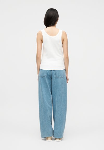 Γυναίκα με μακριά σκούρα μαλλιά, φοράει λευκό αμάνικο τοπ, ανοιχτό μπλε τζιν φαρδιάς γραμμής και μπεζ αθλητικά παπούτσια, γυρισμένη πλάτη σε απλή φόντο.