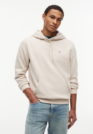 Νεαρός άνδρας που φοράει ανοιχτό μπεζ φούτερ με κουκούλα και ανοιχτό μπλε τζιν, στέκεται με το ένα χέρι στην τσέπη μπροστά σε λευκό φόντο.