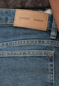 Modré džínsy z denimovej tkaniny s koženou nášivkou na páse, embosovanou s nápisom "SAMSØE SAMSØE". Majú klasický dizajn s piatimi vreckami.