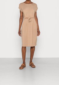 Frau in einem knielangen beigen Kleid mit weißen horizontalen Streifen und einem gebundenen Taillengürtel, kombiniert mit braunen flachen Sandalen.