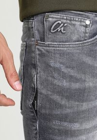 Jeans de mezclilla gris con una textura desgastada, que presentan un logo bordado en el bolsillo trasero y una etiqueta en la costura lateral. Primer plano de una mano.