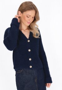 Cardigan blu navy, testurizzato, con scollatura a V, cinque bottoni dorati lungo la parte frontale e maniche lunghe. Fit cropped con un bordo inferiore leggermente sfilacciato.