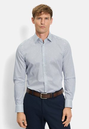 Man draagt een wit overhemd met zwarte stippen, donkerblauwe broek en een bruine leren riem, staand tegen een witte achtergrond.