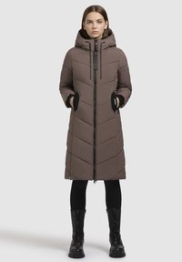 Langer brauner Puffermantel mit Kapuze, ausgestattet mit einem Zickzack-Steppmuster, seitlichen Taschen und einem Reißverschluss vorne. Getragen mit schwarzen Leggings und Stiefeln.