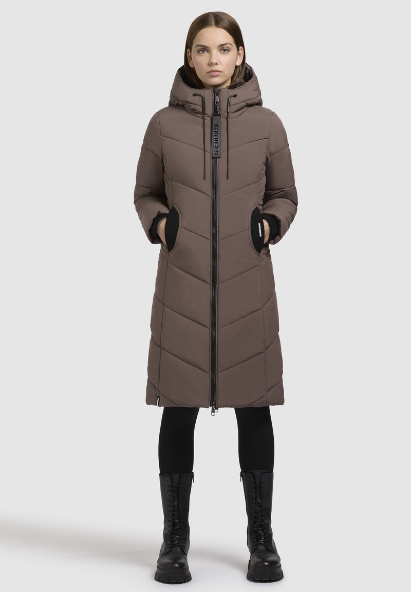 Langer brauner Puffermantel mit Kapuze, ausgestattet mit einem Zickzack-Steppmuster, seitlichen Taschen und einem Reißverschluss vorne. Getragen mit schwarzen Leggings und Stiefeln.