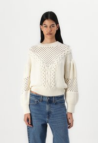 Pull en tricot crème avec un design en câble texturé, des accents en dentelle et des manches bouffantes. Associé à un jean en denim bleu.