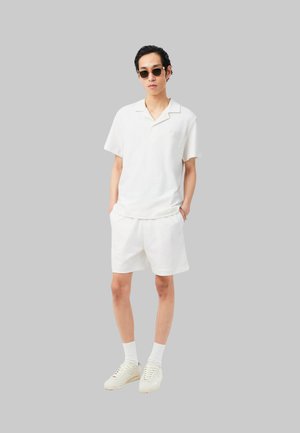 Hombre que lleva una camisa blanca de manga corta, pantalones cortos blancos, calcetines blancos y zapatillas blancas con gafas de sol, de pie frente a un fondo gris liso.