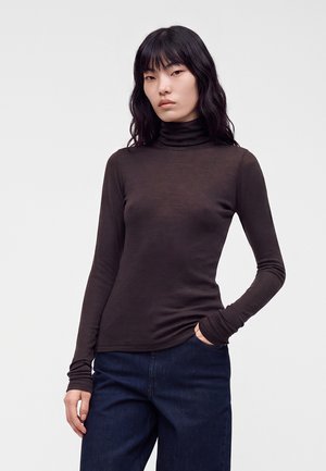 Femme aux longs cheveux noirs portant un col roulé marron foncé ajusté et un jean bleu foncé, debout avec une main dans la poche sur un fond uni.