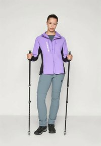 Μωβ και μαύρο εξωτερικό μπουφάν με φερμουάρ και λογότυπο, συνδυασμένο με γκρίζα παντελόνια και μαύρα παπούτσια πεζοπορίας; κρατώντας trekking poles.