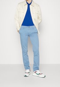 Beige lättviktsjacka, blå piké, ljusblå byxor och vita sneakers med gröna och vinröda detaljer, stående mot en neutral bakgrund.