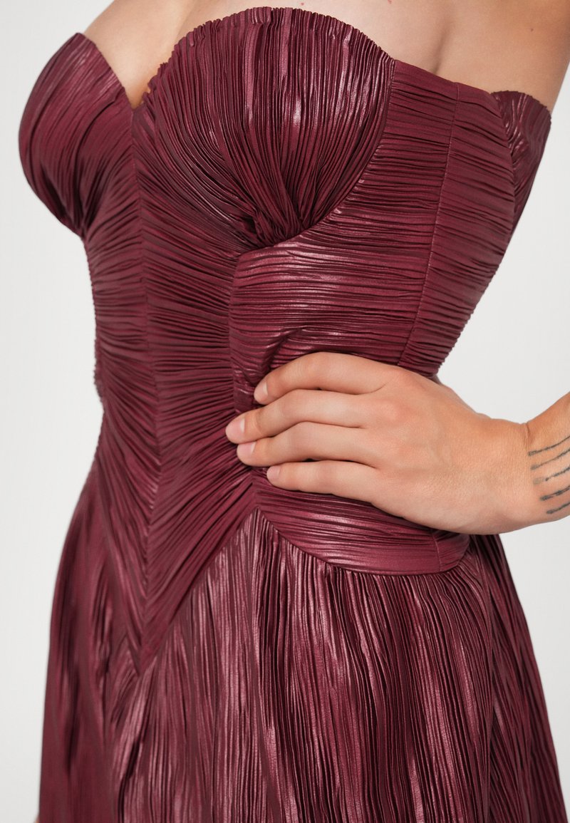 Bordeaux plooienjurk met een strapless halslijn, textuurstof en dartnaden die een getailleerde pasvorm bij de taille en een vloeiende rok creëren.