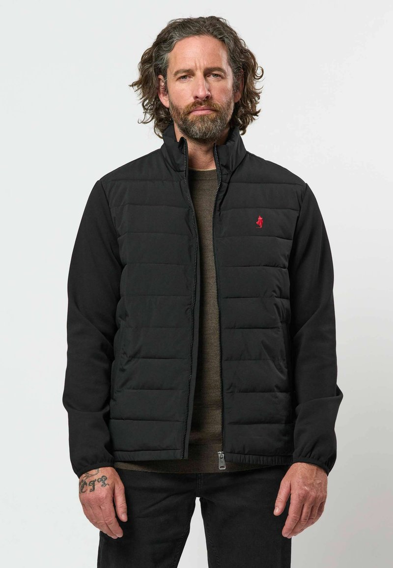 Homme aux cheveux bouclés mi-longs et barbe, portant une veste matelassée noire avec un logo rouge et un pantalon noir sur fond gris.
