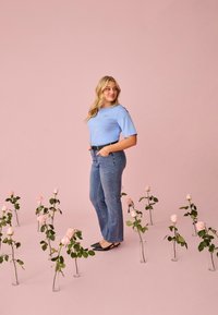 Junge Frau mit blonden Haaren, die ein blaues Hemd und Jeans trägt, steht zwischen pinkfarbenen Rosen in Glasvasen vor einem pinkfarbenen Hintergrund.