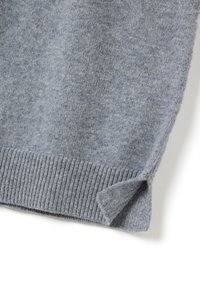 Bordure inférieure d'un pull en laine gris clair avec un ourlet en côtes et une petite fente sur le côté, sur un fond blanc.