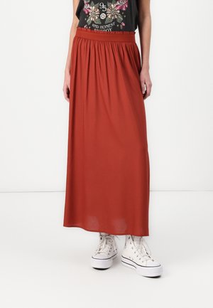 Vrouw draagt een roestkleurige lange rok met een elastische tailleband, witte hoge sneakers en een zwart T-shirt met bloemmotief.