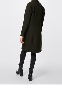 Olijfgroene wollen jas met een gestructureerde silhouet, enkele achterkant split en lange mouwen, gecombineerd met zwarte slim fit broek en laarzen met hak.
