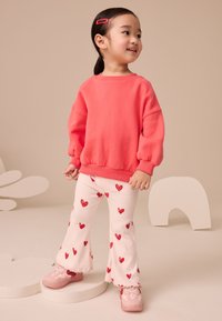 Korálová mikina s nazdobenými rukávmi, v kombinácii s bielymi rebrovanými nohavicami s červenými srdcovými vzormi. Svetloružové tenisky dopĺňajú celý outfit.