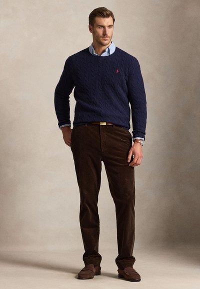 Uomo che indossa un maglione a trecce blu navy sopra una camicia azzurro chiaro con collo, pantaloni marrone scuro e mocassini marroni, in piedi su uno sfondo neutro.
