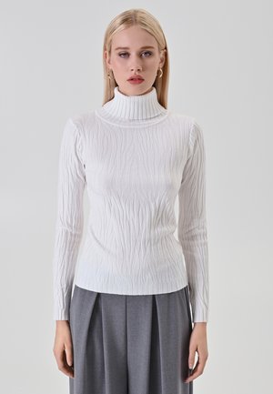 Blond woman wearing white textured turtleneck sweater and gray pleated wide-leg trousers, standing against a plain light background.