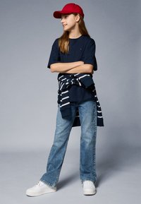 Marineblaues T-Shirt mit Logo, hellblaue Jeans und weiße Sneakers. Rote Kappe mit Schirm und gestreifter Pullover um die Taille gebunden.