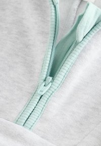 Világosszürke cipzáras kapucnis pulóver menta zöld cipzárral és részletekkel. Puha, texturált anyagból készült, bordázott szegélyű gallérral.