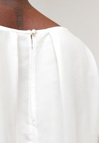 Vit blus med knappstängning i ryggen, gjord av mjukt och slätt tyg samt har en avslappnad, flödande design. Diskret sömdekor.