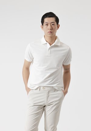 Joven con cabello corto y negro, vestido con una camiseta polo blanca y pantalones beige claro, de pie con las manos en los bolsillos contra un fondo liso.