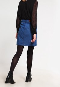 Jupe trapèze en jean avec une teinte bleu clair, une taille ajustée et une poche arrière. Portée avec des collants noirs et des bottines. Haut noir texturé.