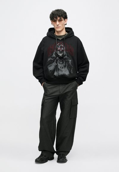 Hanorac negru cu imprimeu grafic, șnururi și buzunar tip kangaroo. Pantaloni cargo negri cu buzunare mari. Tenisi negri și ochelari transparenți.
