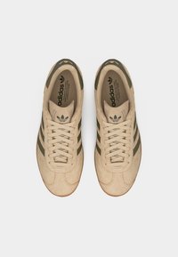 Ζευγάρι μπεζ σουέντ αθλητικά παπούτσια Adidas με ρίγες και κορδόνια σε λαδί πράσινο, από πάνω σε λευκό φόντο.