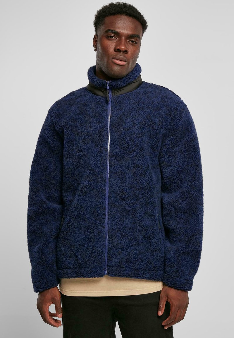 Junger Mann trägt eine marineblaue Reißverschluss-Fleecejacke und ein beiges Hemd, steht vor einem schlichten hellgrauen Hintergrund.