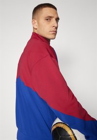 Rød og blå jakke med glidelås og høy krage, med en fargeblokkert design og glatt tekstur, holdt av en person med en sportsball.