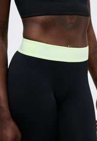 Κοντινό πλάνο ενός ατόμου που φοράει μαύρα ψηλόμεσα κολάν με ανοιχτό κίτρινη ζώνη μέσης με αχνό λογότυπο "NIKE PRO".