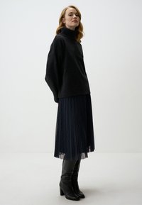 Schwarzer gestrickter Rollkragenpullover, der über einem marineblauen Faltenrock mit einem strukturierten Rautenmuster und kniehohen schwarzen Stiefeln mit Absatz getragen wird.