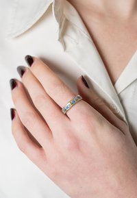 Gold ring featuring a row of blue and green gemstones set into a polished band, worn on a hand with dark nail polish against a white shirt.