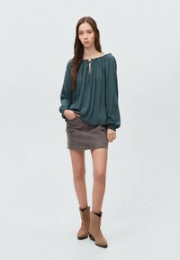 Blusa teal con scollatura arricciata e lacci, abbinata a una gonna in denim grigia con tasche e stivaletti marroni alla caviglia con foderina in pelliccia sintetica.