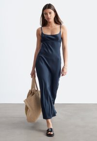 Femme portant une robe bleu foncé sans manches, des sandales noires à bout ouvert, portant un grand sac fourre-tout beige tissé, marchant sur un sol gris.