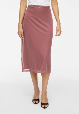 Femme portant une jupe rose poudré longueur genou avec un petit nœud à la taille et des talons ouverts noirs sur fond blanc.
