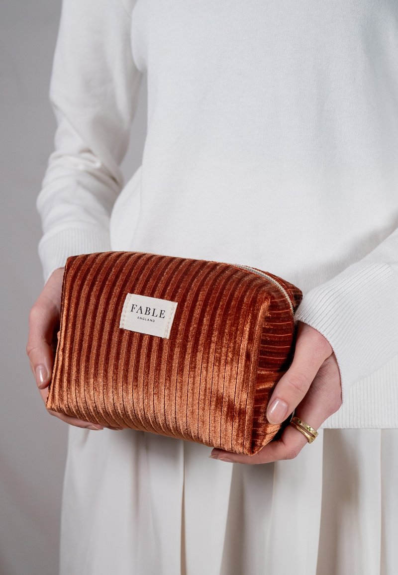 Rust-colored corduroy cosmetic pouch with a textured surface, featuring a white label that reads "FABLE ENGLAND." Zippered top closure.