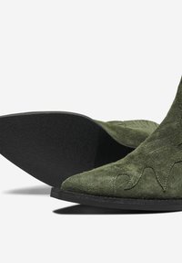 Botoșei de gleznă verzi din suede, cu vârfuri ascuțite, cusături decorative și tălpi din cauciuc texturat negru. Accent pe formă și material.