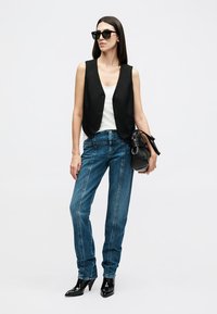 Woman wearing black sleeveless vest, white top, blue jeans, black heeled boots, and sunglasses, holding a black handbag.