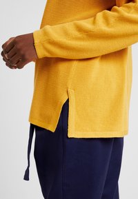 Persona che indossa un maglione giallo a trama con uno spacco laterale e pantaloni blu navy, con le mani incrociate dolcemente davanti.