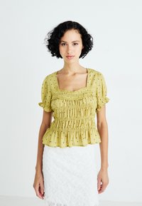 Gele, geplooide blouse met een vierkante halslijn, pofmouwen en kleine bloemrijke versieringen. Gepaard met een witte kant rok.