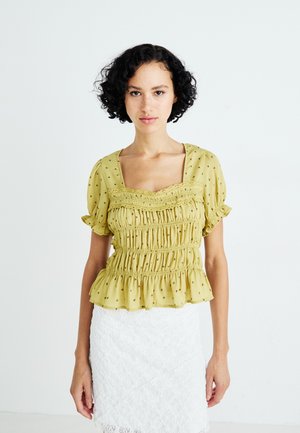 Gele, geplooide blouse met een vierkante halslijn, pofmouwen en kleine bloemrijke versieringen. Gepaard met een witte kant rok.
