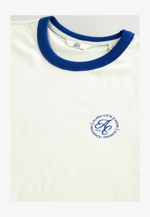 Tricou cu guler rotund din bumbac crem deschis, cu un guler ribatat albastru și un logo circular în albastru închis cu detalii textuale.
