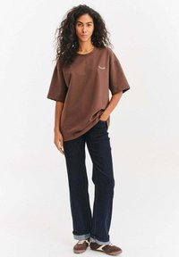 Brązowy oversize'owy t-shirt z okrągłym dekoltem i krótkimi rękawami, z prostym haftowanym detalem. W połączeniu z ciemnoniebieskimi dżinsowymi spodniami.