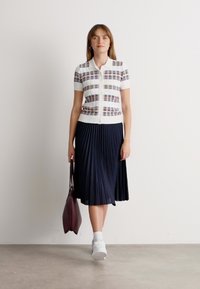 Femme marchant à l'intérieur, portant un haut à rayures à manches courtes et boutonné, une jupe plissée bleu marine, des baskets blanches et portant un sac fourre-tout rouge foncé.