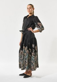 Sort linnedress med blomsterbroderi i pastelfarver, knaplukning foran, lange ærmer, bindebånd i taljen, svajende skirt, parret med sorte slingback-sko.