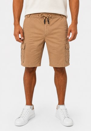 Uomo che indossa pantaloncini cargo beige con coulisse in vita e scarpe da ginnastica bianche, in piedi davanti a uno sfondo uniforme.