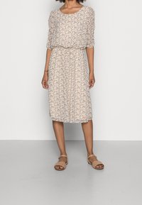 Vrouw in een knie-lengte, witte bloemenjurk met driekwartmouwen en beige open teen sandalen, staande tegen een effen achtergrond.